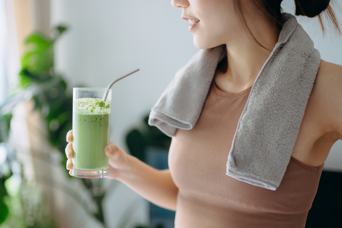 Eine junge Frau steht im Sport Outfit in ihrem Wohnzimmer und hält einen frischen Smoothie in der Hand.