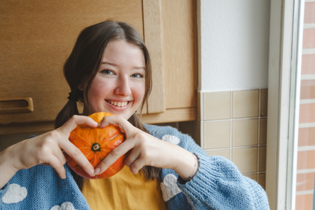 Lächelndes Mädchen hält einen kleinen, orangefarbenen Kürbis und formt ein Herz mit den Händen.