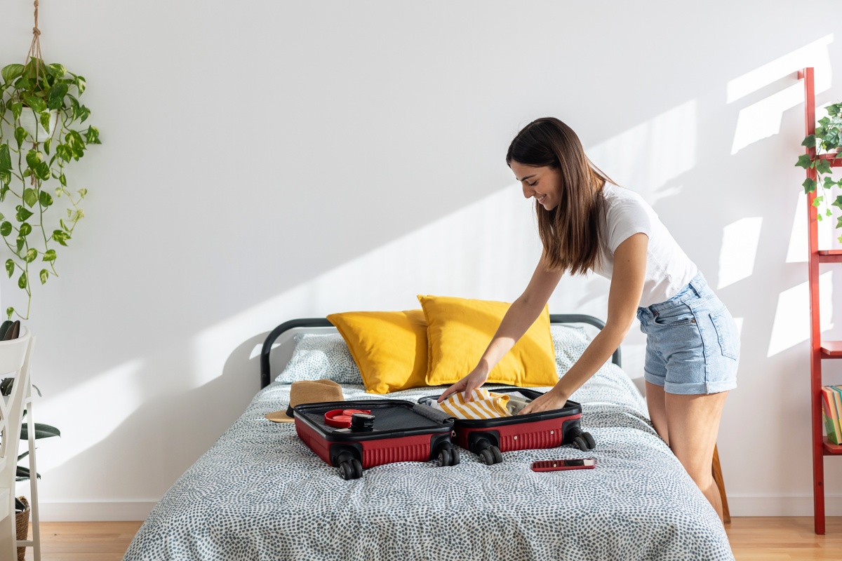 Frau packt einen roten Koffer auf einem Bett mit gelben Kissen in einem hellen, modernen Schlafzimmer.